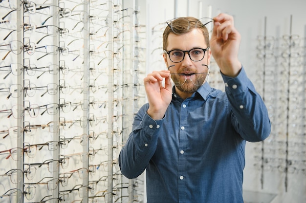 En la tienda de óptica Retrato de un cliente masculino sosteniendo y usando diferentes anteojos eligiendo y probándose nuevos anteojos en la tienda óptica Hombre recogiendo marco para el primer plano de corrección de la visión