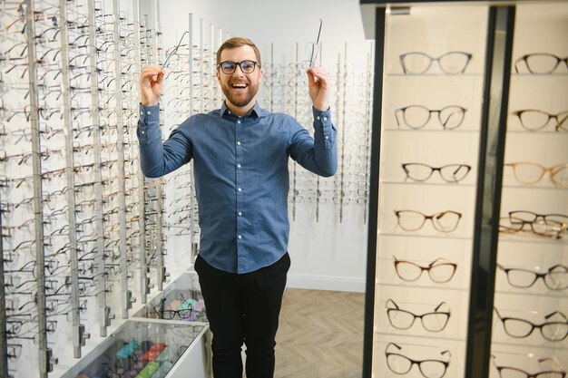 En la tienda de óptica Retrato de un cliente masculino sosteniendo y usando diferentes anteojos eligiendo y probándose nuevos anteojos en la tienda óptica Hombre recogiendo marco para el primer plano de corrección de la visión