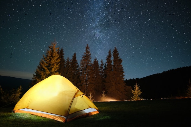 Tienda turística iluminada brillante cerca de la hoguera que brilla intensamente en el camping en las montañas oscuras bajo el cielo nocturno con estrellas brillantes. Concepto de estilo de vida activo.