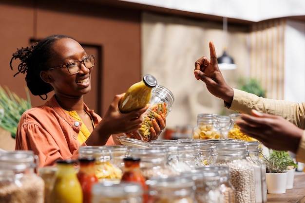 En una tienda, un vendedor afroamericano ofrece alimentos orgánicos producidos regionalmente, verduras frescas y artículos esenciales para la despensa sin plástico son entregados a la cliente femenina por el tendero.