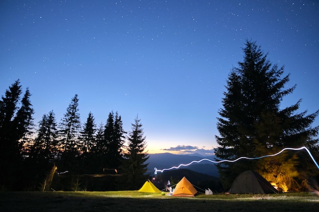 Tiendas de campaña brillantemente iluminadas brillando en el camping en montañas oscuras bajo el cielo cubierto de estrellas de la noche. Estilo de vida activo y concepto de viaje.