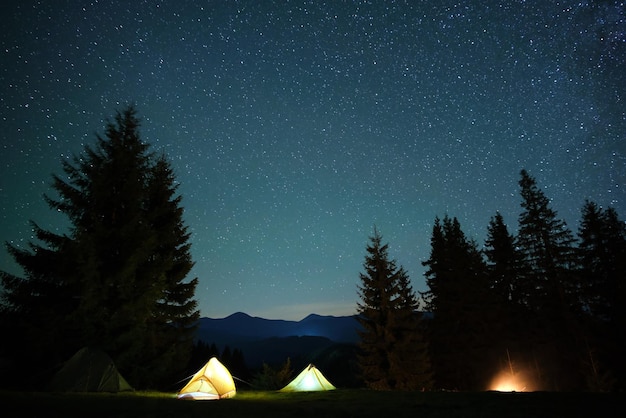 Tiendas de campaña turísticas iluminadas brillantes cerca de una hoguera brillante en un camping en un bosque de montaña oscuro bajo un cielo nocturno con estrellas brillantes. Estilo de vida activo y concepto de vida al aire libre.