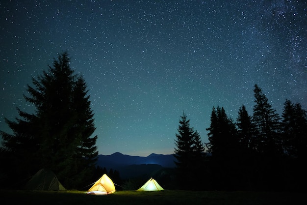 Tiendas de campaña turísticas iluminadas que brillan intensamente en el camping en montañas oscuras bajo el cielo nocturno con estrellas brillantes Concepto de estilo de vida activo