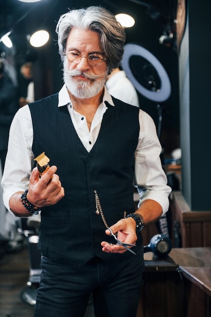 Tiene equipo de barbería Elegante hombre senior moderno con cabello gris y barba está en el interior