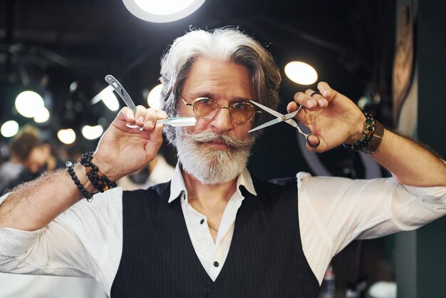 Tiene equipo de barbería Elegante hombre senior moderno con cabello gris y barba está en el interior