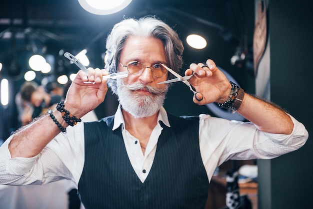 Tiene equipo de barbería Elegante hombre senior moderno con cabello gris y barba está en el interior