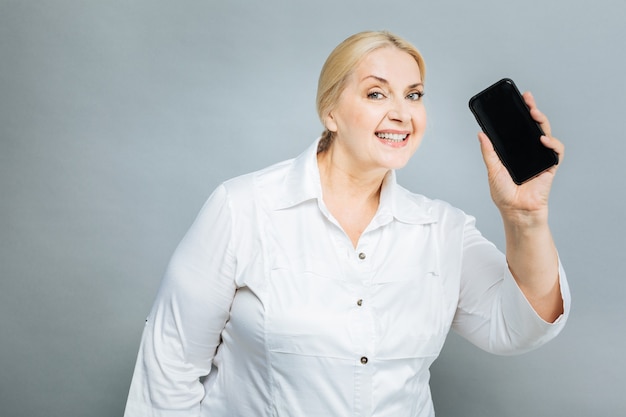 Tienes una llamada. Mujer encantada positiva demostrando teléfono y manteniendo la sonrisa en la cara mientras está de pie aislado en gris