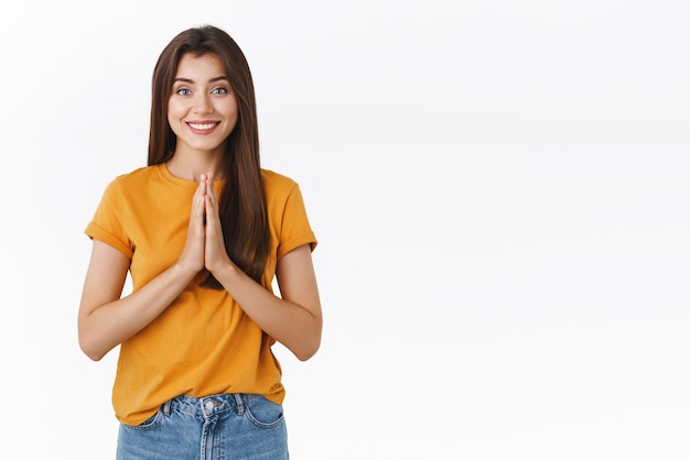 Tierna, amable y feliz mujer joven con expresión de ángel confiable se toma de la mano en un gesto de súplica de oración sonriendo pidiendo favor rogando o agradeciendo ayuda de fondo blanco