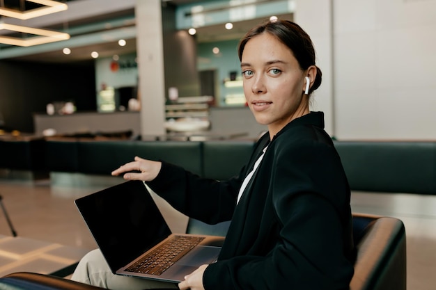 Tierna dama bonita con cabello oscuro vestido con una chaqueta oscura en los auriculares sostiene una computadora portátil y trabaja en la oficina en un día soleado