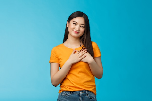 Tierna y encantadora chica asiática de aspecto amigable con corte de pelo corto oscuro, toque el pecho, presione las palmas del corazón, mire con admiración y ternura, sonriendo alegremente, contemple la conmovedora escena, fondo azul.