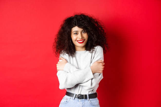 Tierna joven abrazándose a sí misma, sintiéndose cómoda y feliz, sonriendo tontamente a la cámara, de pie contra el fondo rojo.