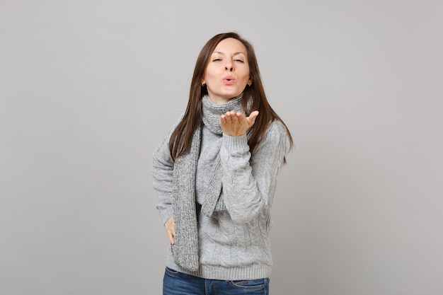 Tierna joven en bufanda de suéter gris que sopla enviando beso de aire aislado sobre fondo de pared gris en estudio. Gente de estilo de vida de moda saludable emociones sinceras concepto de estación fría. Simulacros de espacio de copia.