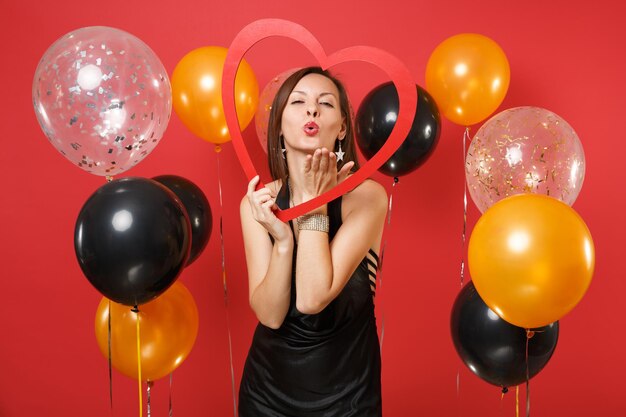 Tierna joven vestida de negro con gran corazón de madera rojo, labios que soplan, enviando beso de aire en globos de aire de fondo rojo. Día de San Valentín, concepto de fiesta de vacaciones de maqueta de cumpleaños de feliz año nuevo.