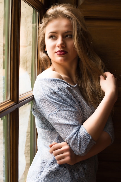 Tierna modelo rubia con maquillaje profesional y cabello largo posando en la ventana