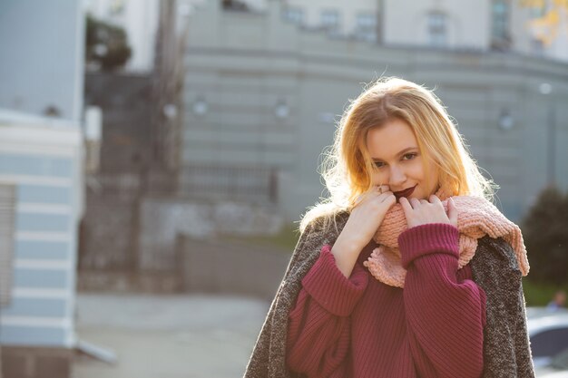 Tierna mujer rubia vestida con abrigo y bufanda, posando con luz de fondo suave. Espacio para texto