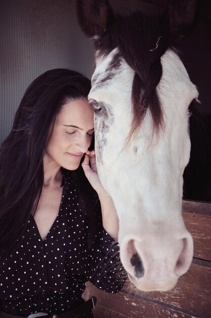 Tierna mujer vestida de negro con los ojos cerrados acariciando un caballo blanco parado en un puesto con valla de madera en una zona rural