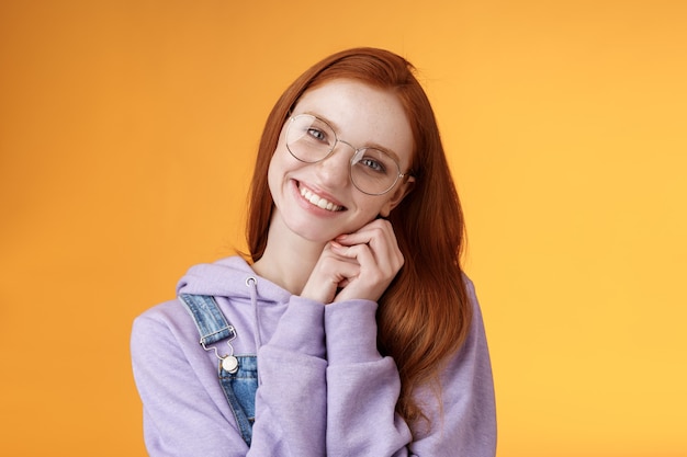 Tierna niña de jengibre femenina ojos azules con gafas sudadera con capucha palmas delgadas sonriendo recibir dulce lindo regalo creo que eres adorable amiga querida agradecida sonriendo alegremente difundir amor, fondo naranja.