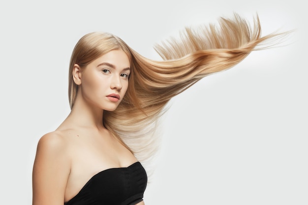 Tierno. Hermosa modelo con cabello rubio largo, liso y volador sobre fondo blanco de estudio. Modelo caucásico joven con piel bien cuidada y cabello soplando en el aire. Concepto de cuidado de salón, belleza, moda.