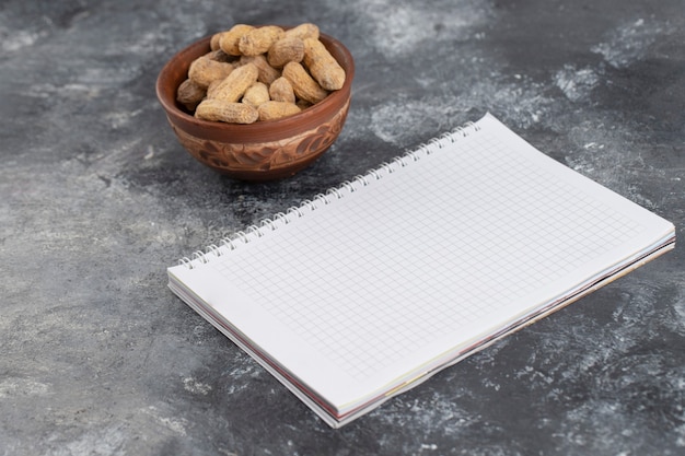 Tigela cheia de amendoim frito com casca com caderno aberto colocado sobre a mesa de pedra.