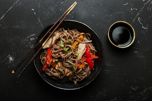 Tigela com frango asiático frito macarrão soba com vegetais em um fundo de pedra preto escuro de cima, prato de macarrão chinês tailandês ou japonês com molho de soja