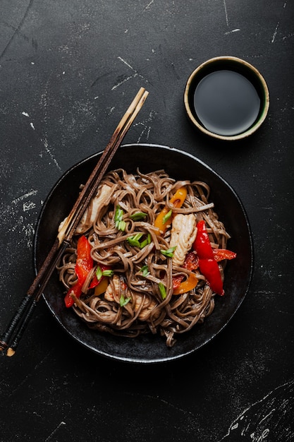 Tigela com frango asiático frito macarrão soba com vegetais em um fundo de pedra preto escuro de cima, prato de macarrão chinês tailandês ou japonês com molho de soja