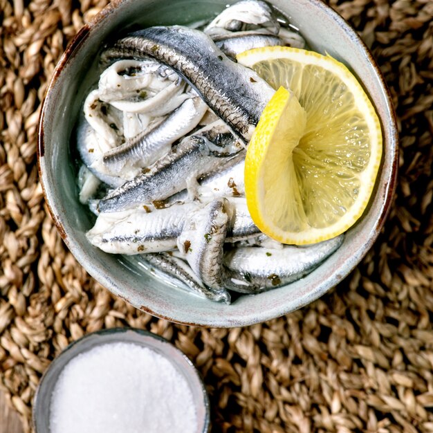 Tigela de anchovas em conserva ou filé de sardinha em óleo, servido com limão, azeite, sal para tapas ou antepastos sobre fundo de madeira velho. Postura plana, copie o espaço. Imagem quadrada