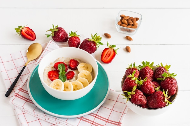 Tigela de aveia com banana e morango, copo de leite em uma mesa branca. alimentos orgânicos saudáveis.