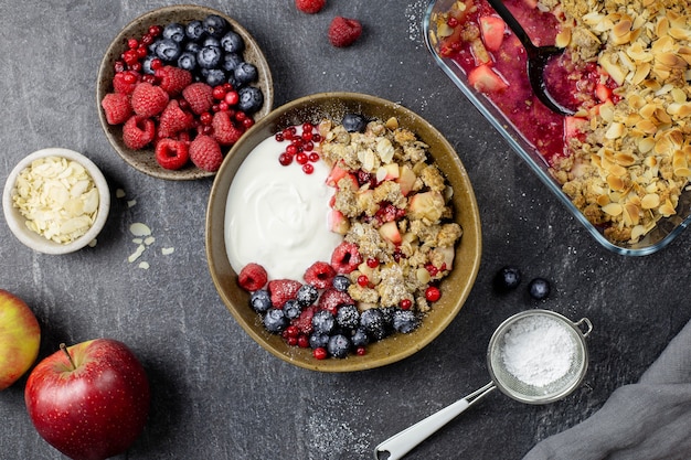 Tigela de granola caseira com iogurte e frutas frescas no fundo escuro de concreto de pedra. crumble de amora e maçã crocante com flocos de amêndoa e aveia.