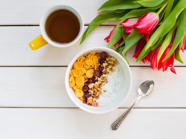 Tigela de iogurte delicioso com flocos de milho, nozes e geleia em uma mesa de madeira branca. conceito de nutrição saudável e orgânica. tulipas com uma xícara de chá e café da manhã. vista superior, copie o espaço