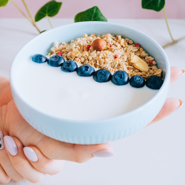 Foto tigela de pequeno-almoço saudável com muesli e iogurte nas mãos de mulher em fundo rosa e mármore