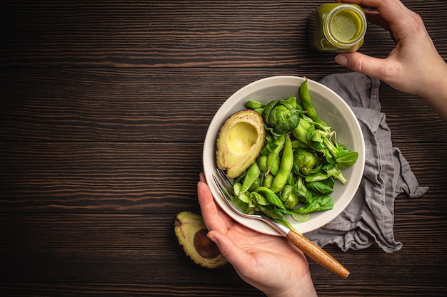 Tigela de salada saudável nas mãos femininas de cima e smoothie de desintoxicação verde em uma garrafa com fundo de madeira, vista superior. Mulher comendo uma refeição vegana saborosa, dieta de alimentos crus e conceito de alimentação limpa, copie o espaço