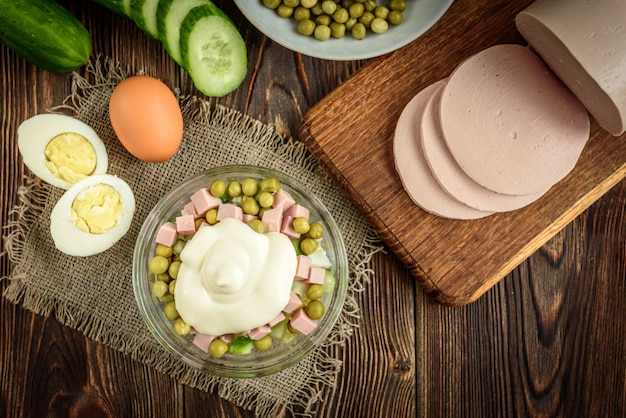 Tigela de salada tradicional russa "Olivier" e ingredientes: salsicha cozida, pepino, ovos, ervilhas e maionese na mesa de madeira escura.