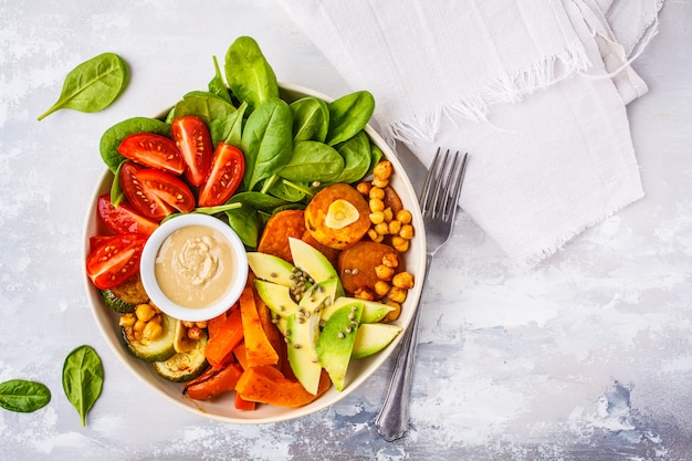 Tigela de salada vegano com legumes cozidos, grão de bico, abacate e molho de tahine em um fundo branco