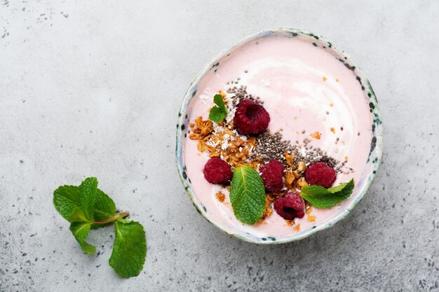 Tigela de smoothie de banana e framboesa rosa com granola, sementes de chia, coco e hortelã em concreto cinza claro.