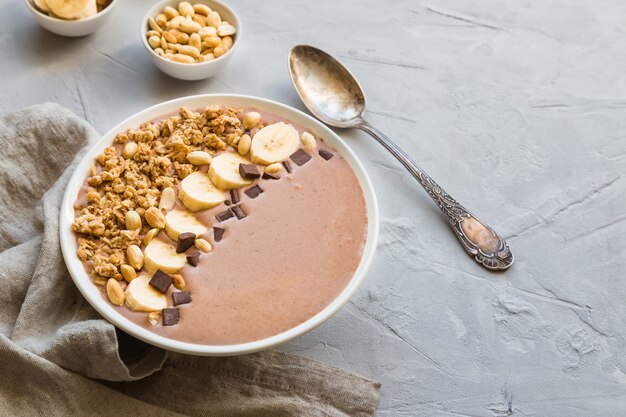 Tigela de smoothie de chocolate com bananas, granola e amendoim em fundo de concreto cinza claro. Café da manhã vegetariano saudável.