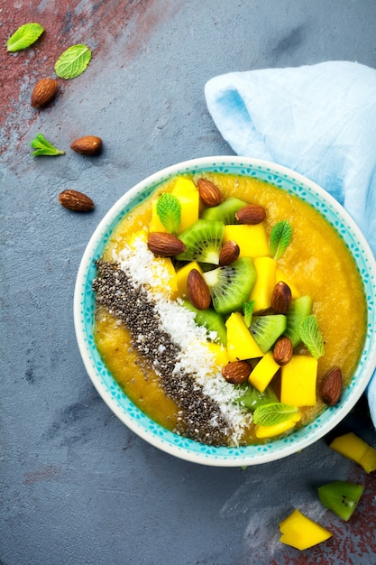 Tigela de suco de manga com pedaços de kiwi, manga, raspas de coco e sementes de chia em uma pedra