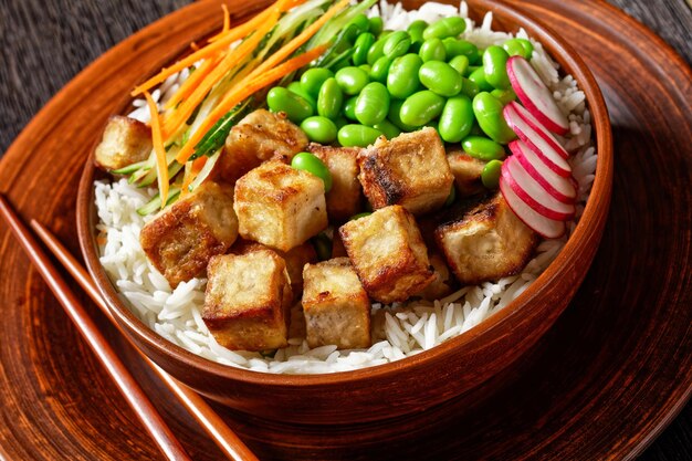 Tigela de tofu vegano com arroz basmati feijão edamame rabanete em fatias finas de pepino e salada de cenoura em uma tigela de barro com pauzinhos vista horizontal de cima closeup