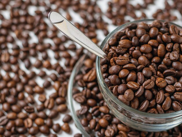 Tigela de vidro com grãos de café e colher de metal na mesa branca com grãos de café espalhados.