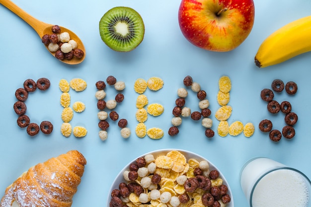 Tigela e colher com bolas de chocolate secas, anéis, flocos de milho, copo de leite, croissant e frutas maduras frescas no café da manhã saudável de cereais de fibra. conceito de cereais