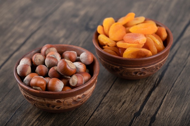 Tigelas cheias de avelãs saudáveis e frutas secas de damasco colocadas sobre uma mesa de madeira.