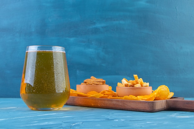 Tigelas de pão torrado e batatas fritas em uma placa ao lado da caneca de cerveja, sobre o fundo azul.