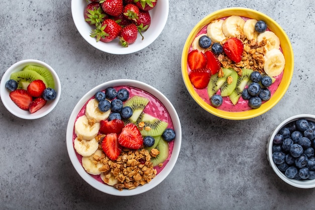 Tigelas de smoothie de açaí de verão com morangos, banana, mirtilos, kiwis e granola em fundo cinza de concreto. taça de café da manhã com frutas e cereais, close-up, vista de cima, comida saudável