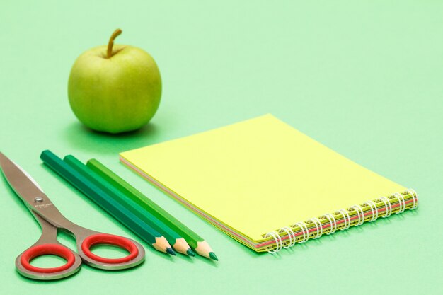 Tijeras, lápices de colores, cuaderno y manzana sobre fondo verde.