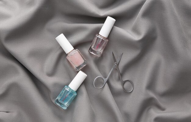 Tijeras de manicura y botellas de esmalte de uñas sobre un fondo de seda gris Concepto de belleza