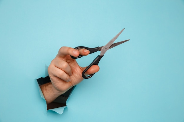 Foto tijeras en mano masculina de un agujero rasgado en papel azul.