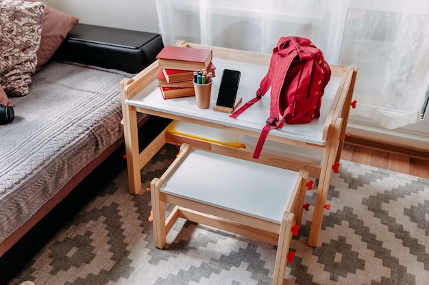 Útiles escolares en el escritorio de la escuela. Mochila roja, auriculares blancos, cuaderno, grandes libros rojos, bolígrafos en el frasco yacían sobre el pupitre blanco.