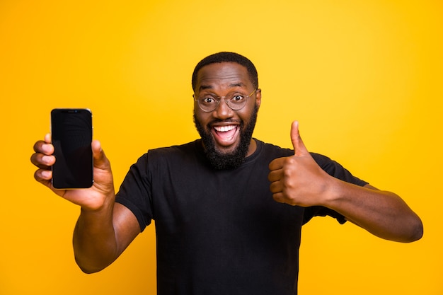 Ótima promoção, escolha! Retrato de um homem afro-americano engraçado segurar smartphone mostrar polegar para cima sinal aconselhar selecionar dispositivo sugerir escolher usar camiseta elegante parede amarela isolada