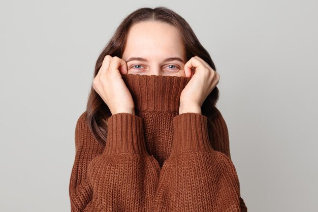 tímida mujer de cabello marrón graciosa con un suéter marrón de pie aislada sobre un fondo gris claro escondiendo la mitad de su cara con el cuello del suéter mirando a la cámara con ojos felices