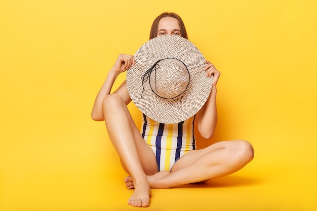 Tímida mujer sonriente con traje de baño a rayas aislado de fondo amarillo escondiendo su rostro con su sombrero de vacaciones tropicales de verano