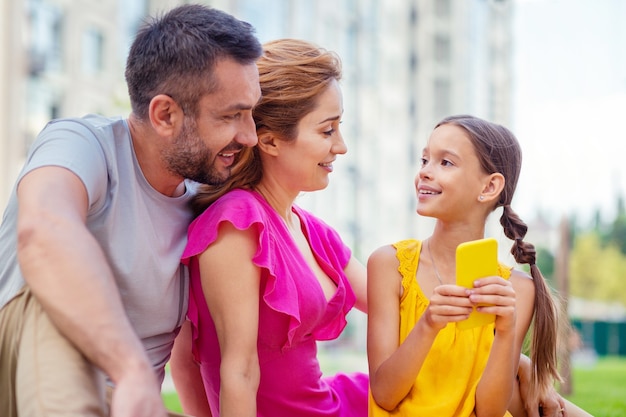 Ótimo humor. Menina alegre e positiva olhando para os pais, segurando um smartphone na mão